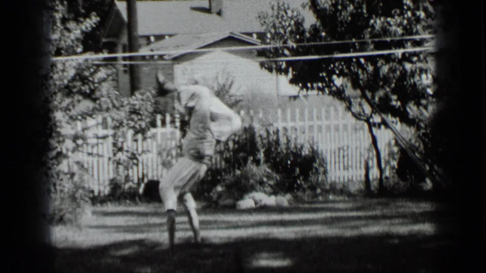 In 1939 New York home-movie footage, a man holds a handstand in a sunny residential backyard near shrubs and a white fence.