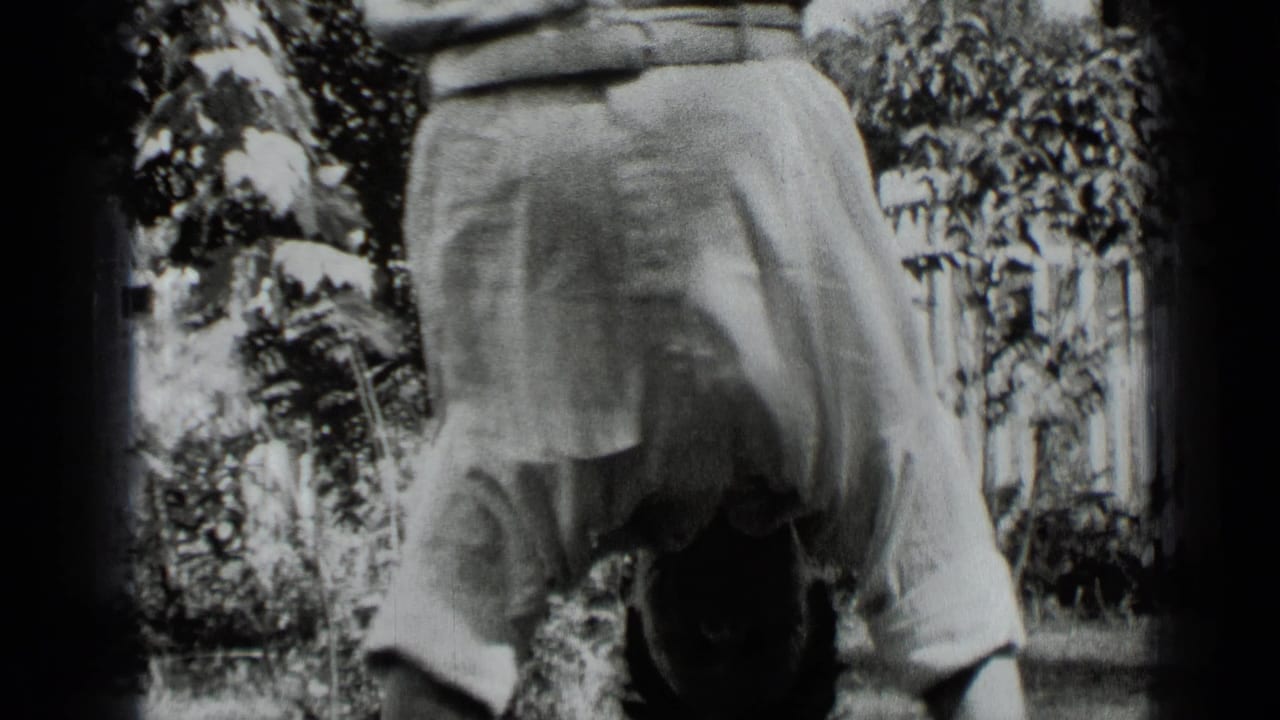 The home-movie frame shows the man upside down in light trousers, with garden foliage and a picket fence behind him.