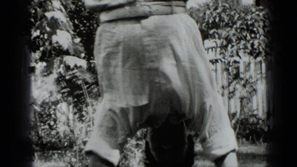 The home-movie frame shows the man upside down in light trousers, with garden foliage and a picket fence behind him.