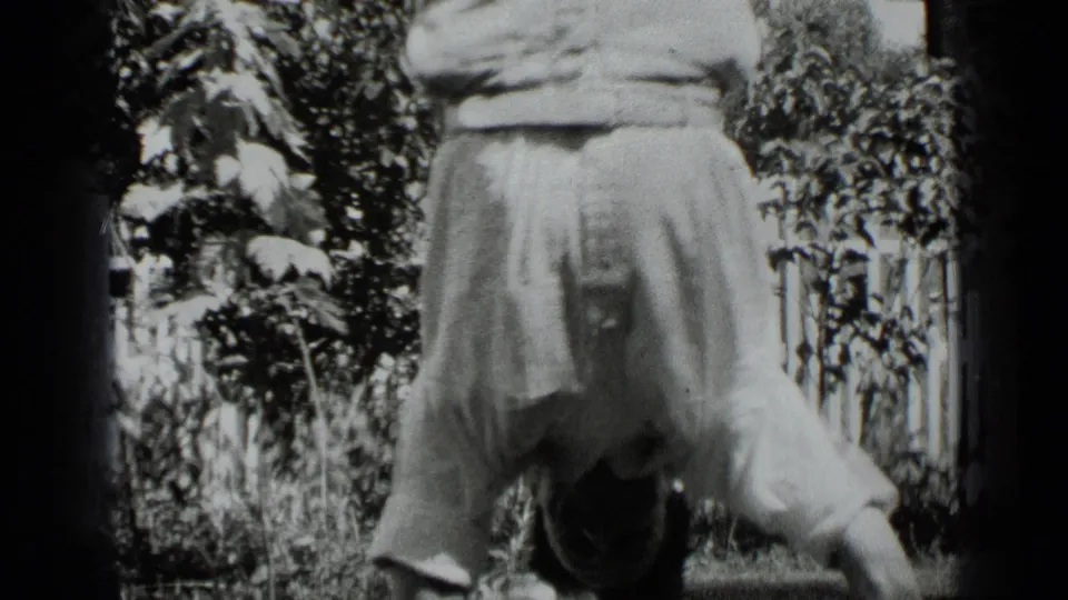 A closer 8mm view shows the man inverted during the handstand, with the backyard fence and leafy plants in the background.