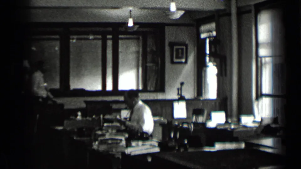 The final frame shifts to an interior room where seated figures appear near desks, windows, and wall hangings in black-and-white 8mm footage.