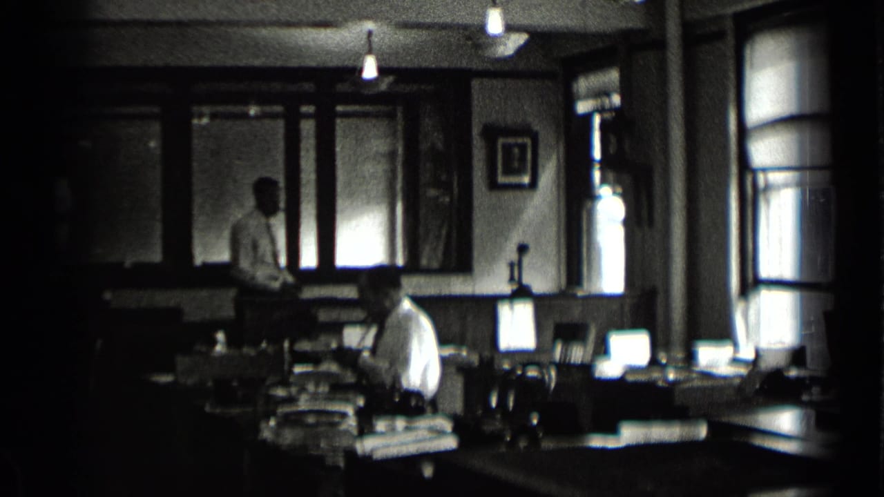 Dim office with men in shirts and ties working among desks, papers, windows, and lamps.