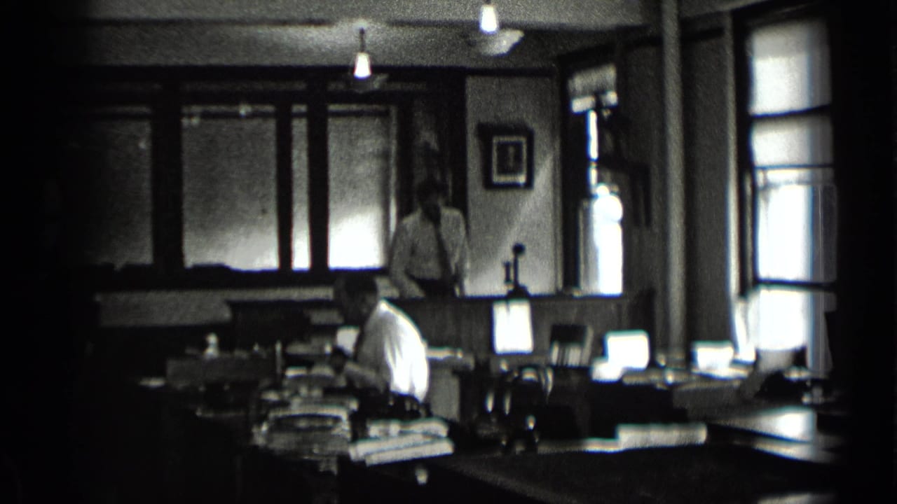 1939 New York office frame showing a seated worker at a desk and another man standing near back windows in low light.