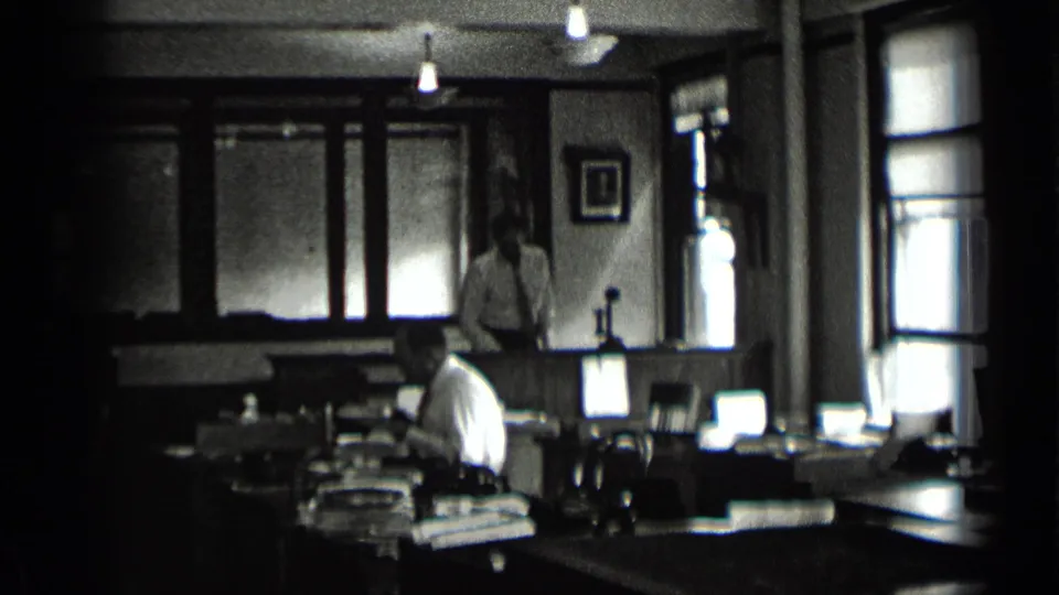 1939 New York office frame showing a seated worker at a desk and another man standing near back windows in low light.