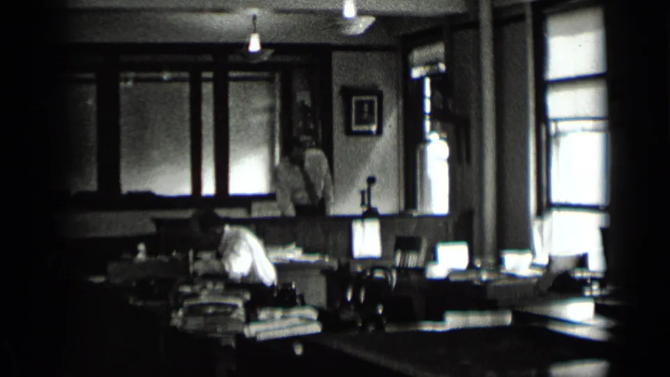 1939 New York office interior with men moving and working among paperwork, desks, lamps, and bright side windows.