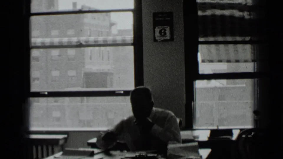 1939 New York office view with a silhouetted seated man near large windows, desk items, and a small wall poster.