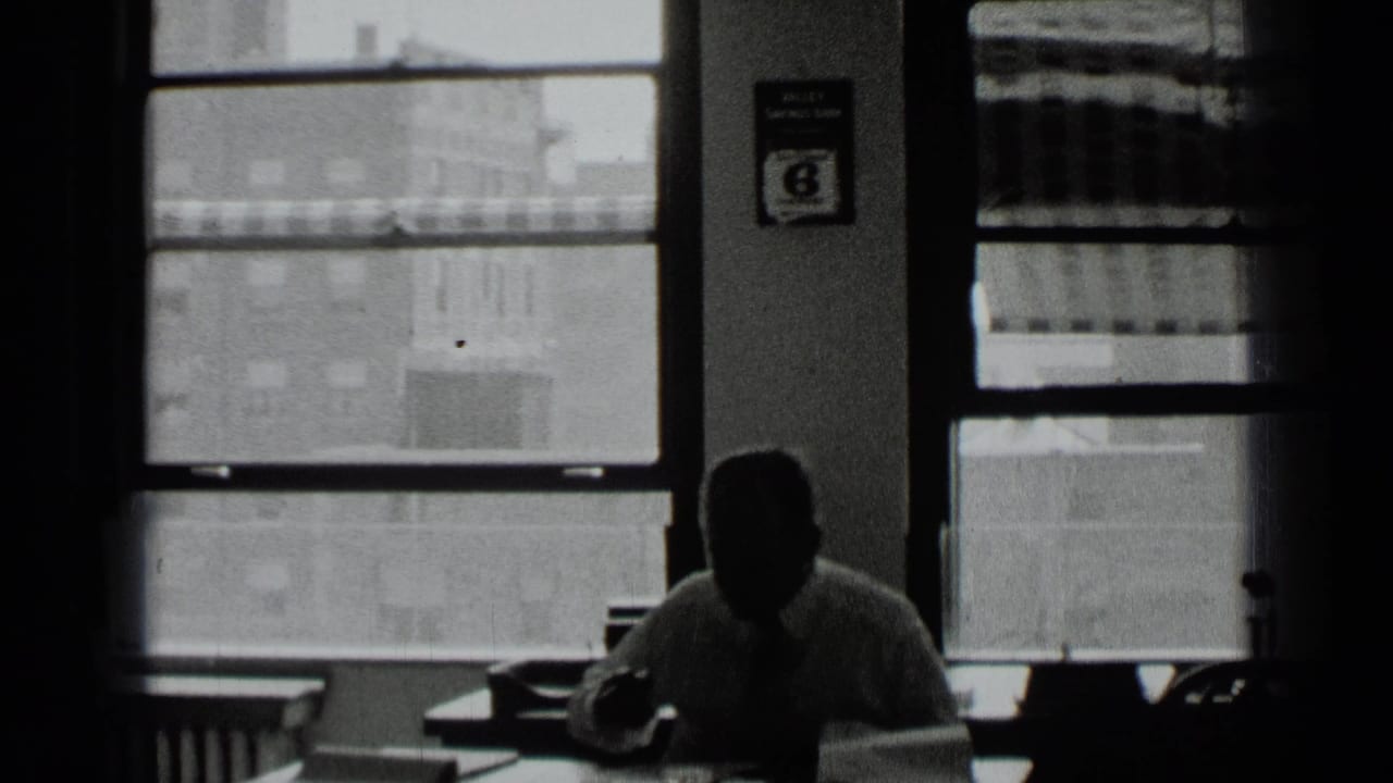 Man seated at a desk in a dim office with large windows behind him.