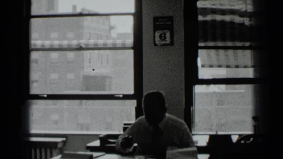 In 1939 New York home-movie footage, a man sits at a desk in a shadowed office while city buildings show through large windows.