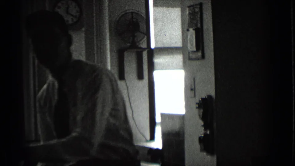 A man in a light shirt and tie stands inside a dim New York office, with a wall clock, wall fan, and doorway visible behind him.