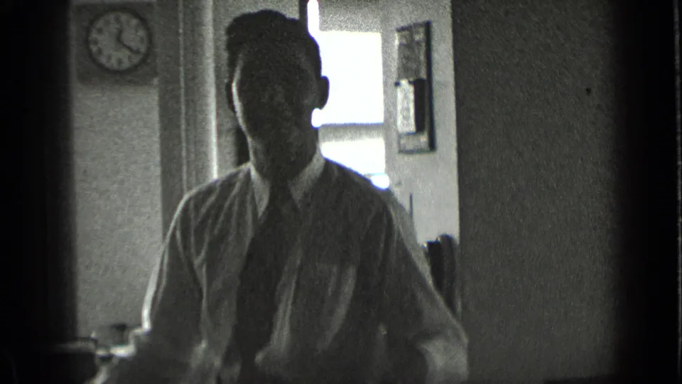 In the final office frame, the man faces the camera in partial light, with a wall clock, bright window, and framed wall item behind him.