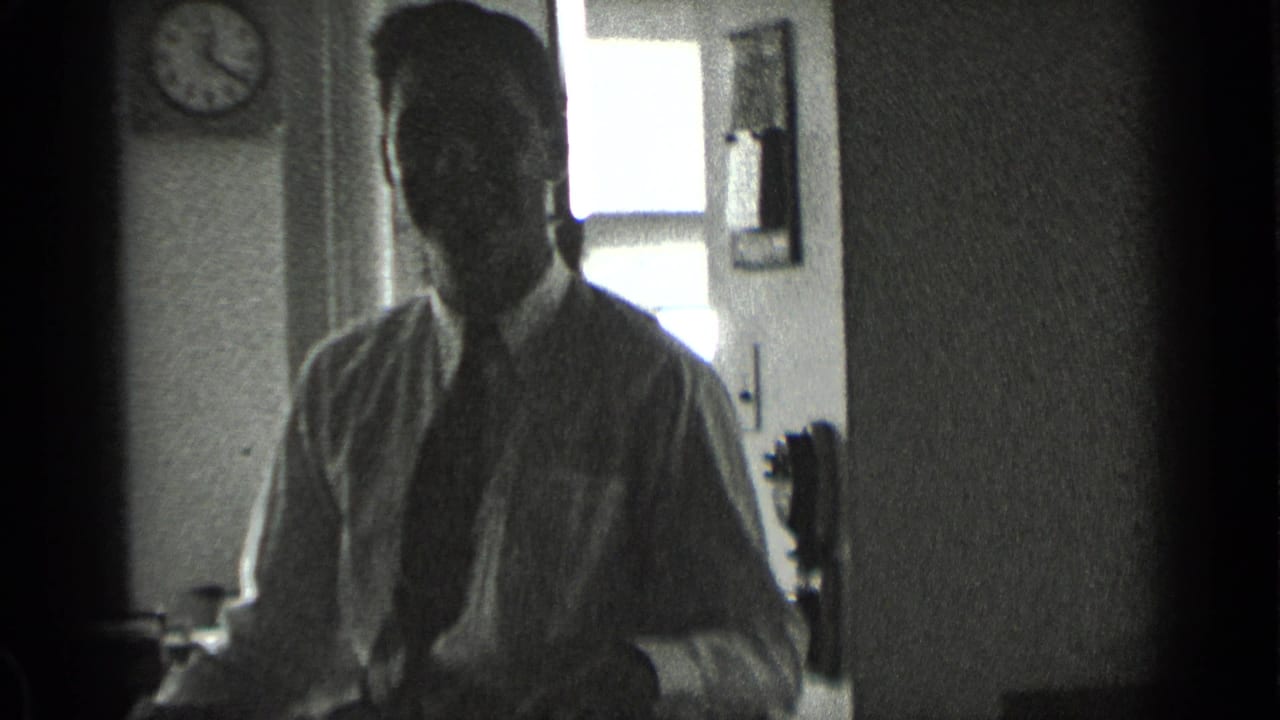 Man in a white shirt and tie sits near a bright window in a dim interior.