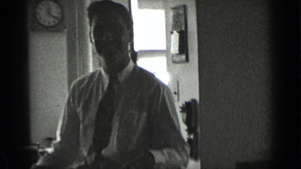 In 1939 New York home-movie footage, a man in a white shirt and tie sits indoors near a bright window and appears to face the camera.