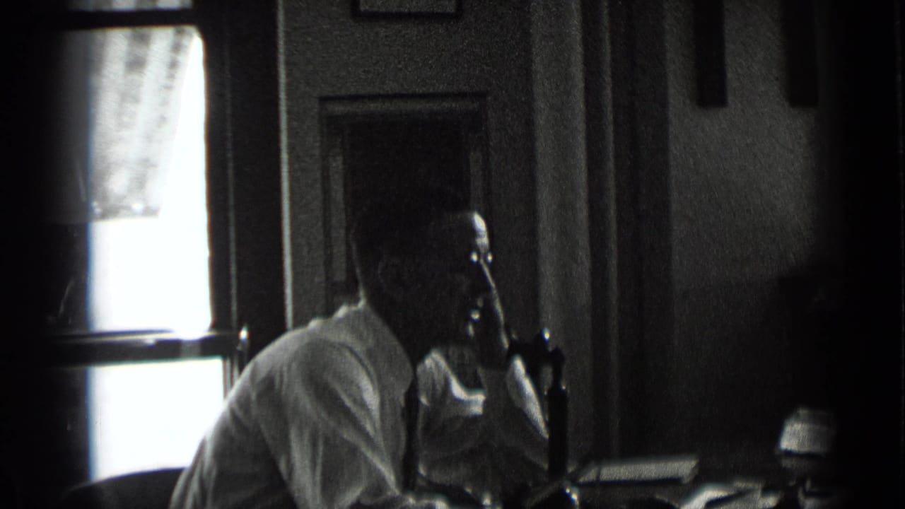 In 1939 New York footage, the man sits in profile at a desk with a telephone receiver to his ear, lit from a bright window behind him.