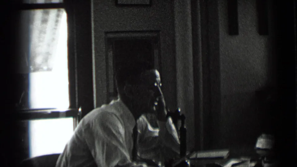 In 1939 New York footage, the man sits in profile at a desk with a telephone receiver to his ear, lit from a bright window behind him.