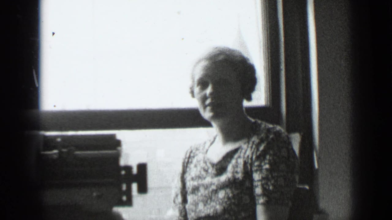 Woman in a patterned dress stands near a bright window with a typewriter in the foreground.
