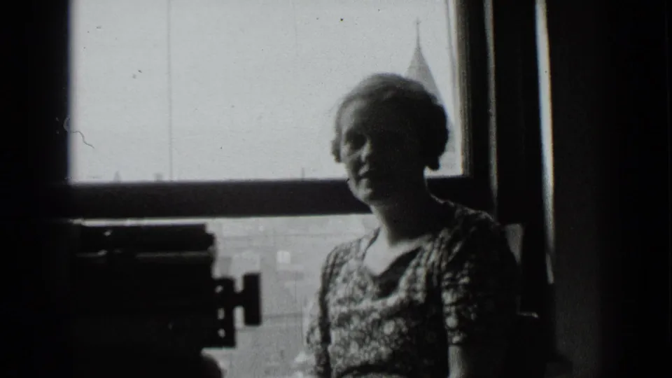 A backlit woman stands beside a window in a New York interior, with a typewriter visible in the shadowed foreground.