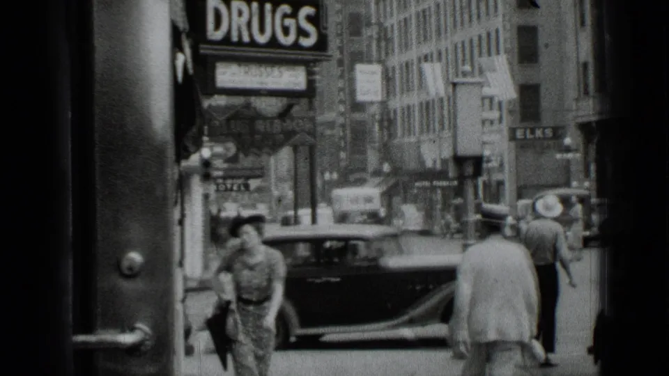 A 1939 New York street view shows pedestrians passing a classic car beneath storefront signage including a large DRUGS sign.