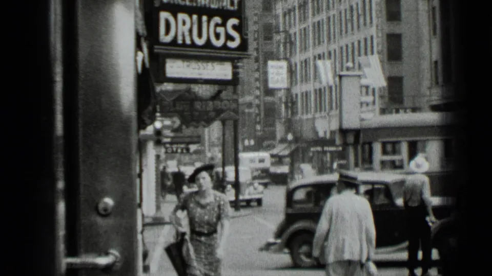 1939 New York street scene with a woman walking toward camera beneath a large DRUGS sign as pedestrians and automobiles fill the background.