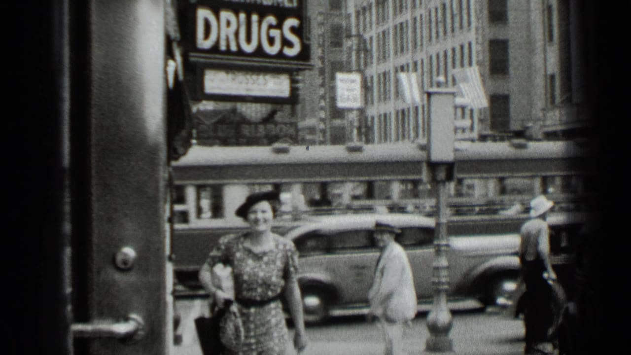 A woman in a patterned dress and dark hat walks along a New York sidewalk in 1939, with traffic and the DRUGS sign visible behind her.