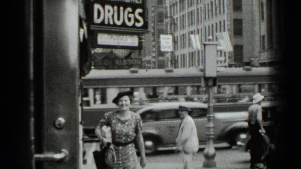 A woman in a patterned dress and dark hat walks along a New York sidewalk in 1939, with traffic and the DRUGS sign visible behind her.