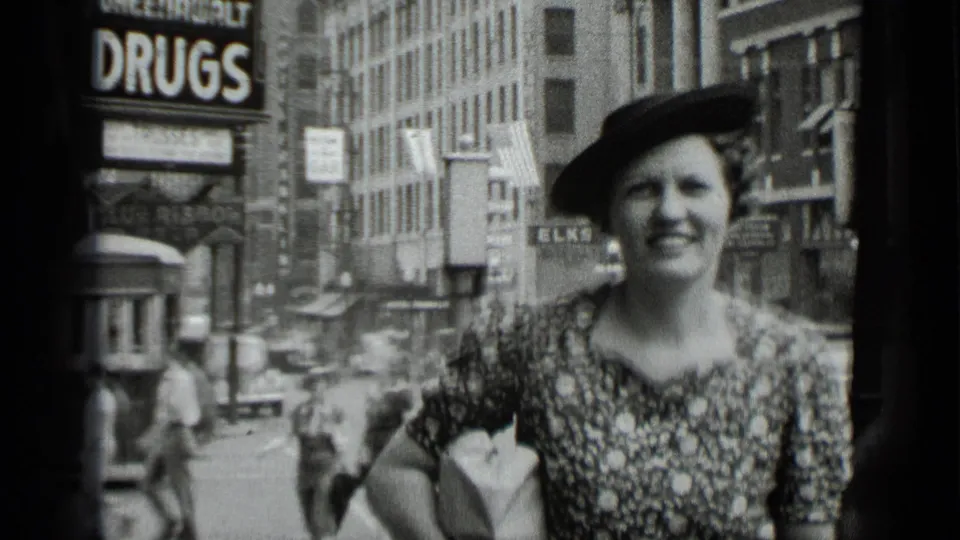 Close 1939 New York home-movie view of a smiling woman in a patterned dress and hat, framed against storefronts, pedestrians, and street traffic.