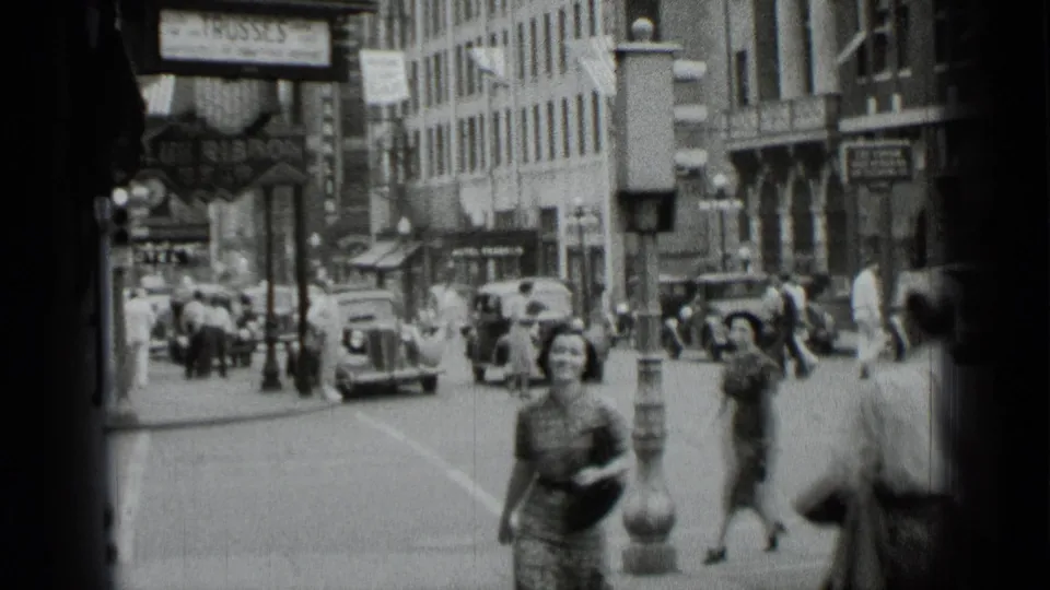 Wide 1939 New York street view showing the woman farther down the sidewalk, pedestrians, vintage automobiles, commercial buildings, and drug store signage.