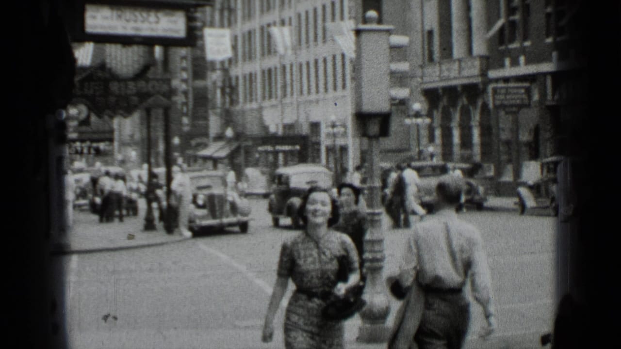 Pedestrians walk along a busy city sidewalk with cars and storefront signs behind them.