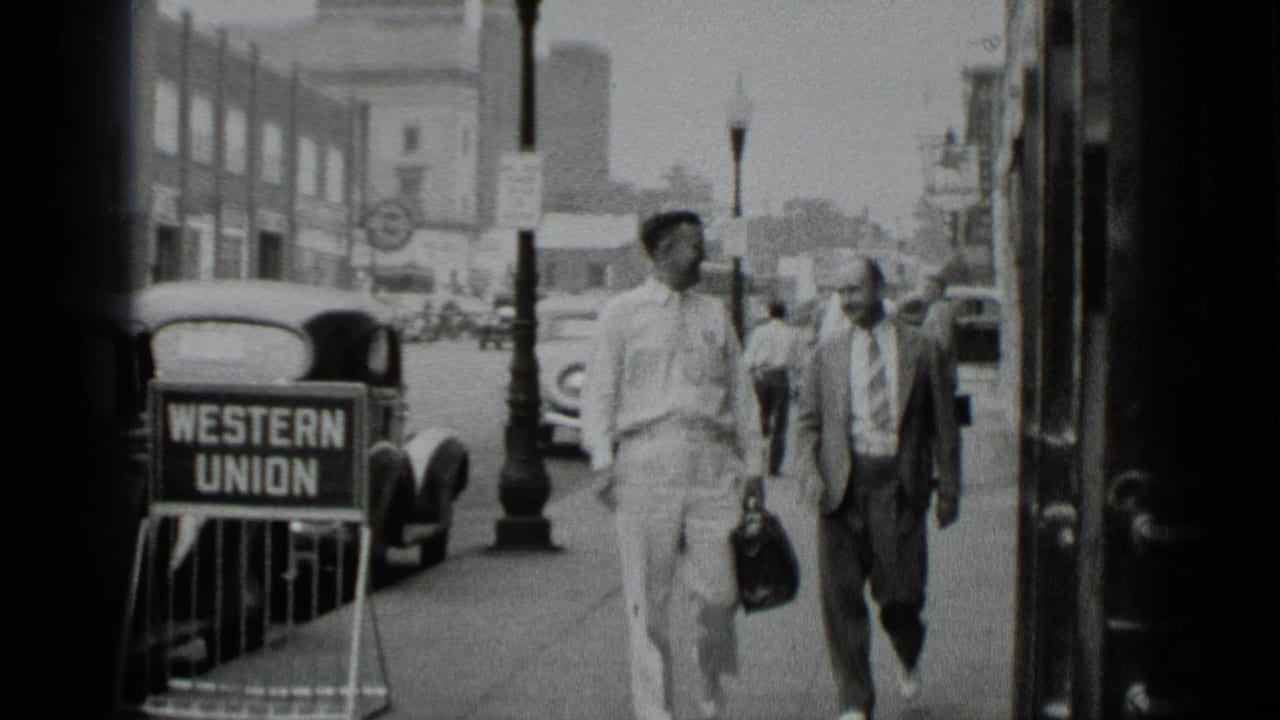 Two men in light shirts and suits walk along a 1939 New York sidewalk beside a visible Western Union sign and period cars.