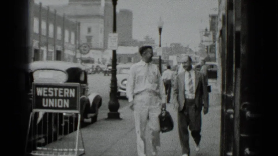 Two men in light shirts and suits walk along a 1939 New York sidewalk beside a visible Western Union sign and period cars.
