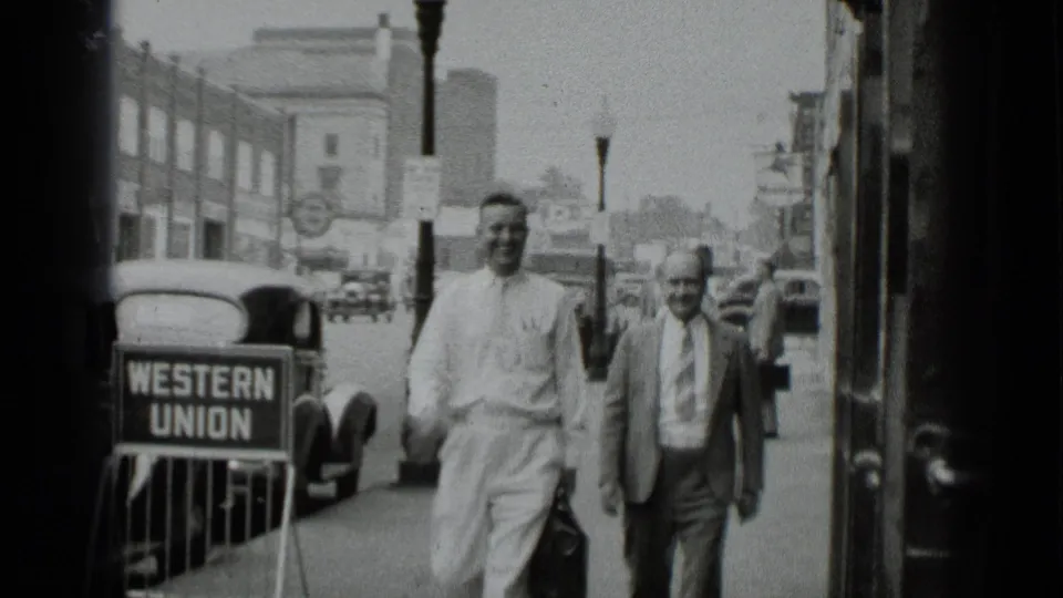 A 1939 New York sidewalk view shows two men walking toward the camera near a Western Union sign and rounded automobiles.