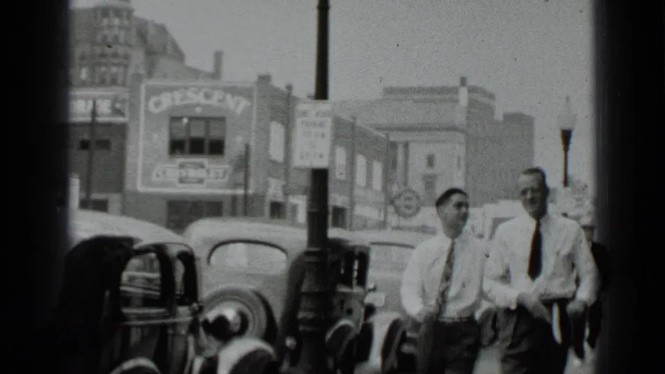 1939 New York street frame with two men on the sidewalk, period cars, storefronts, and a Crescent Chevrolet sign in the background.