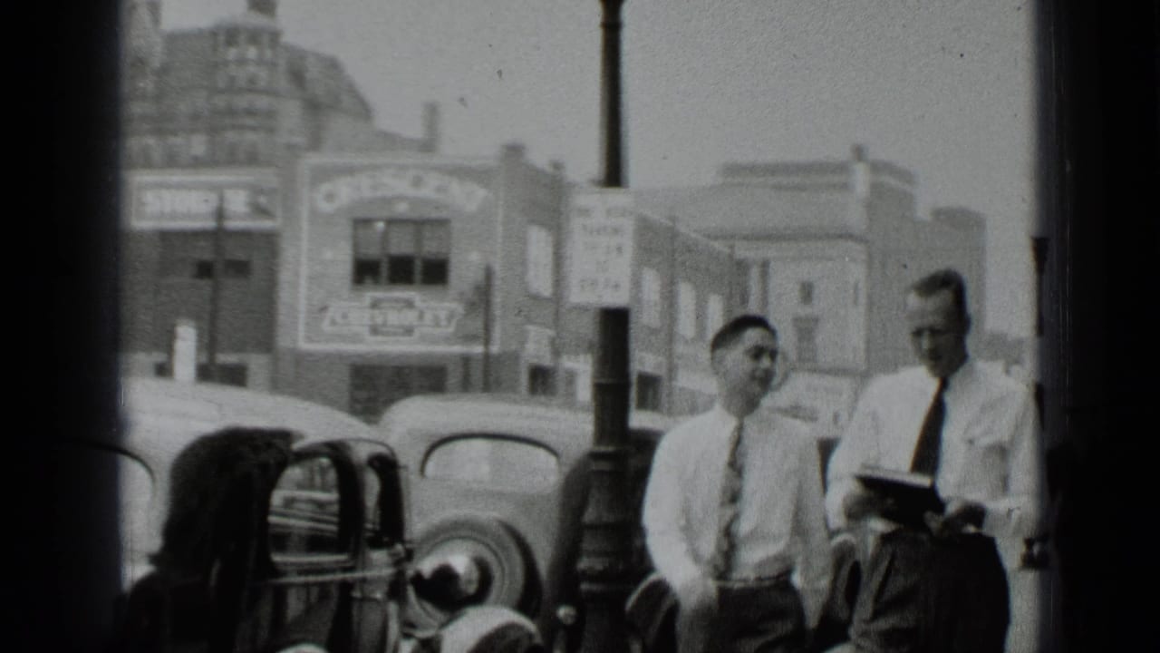 Two men in shirts and ties walk beside parked cars on a city street.