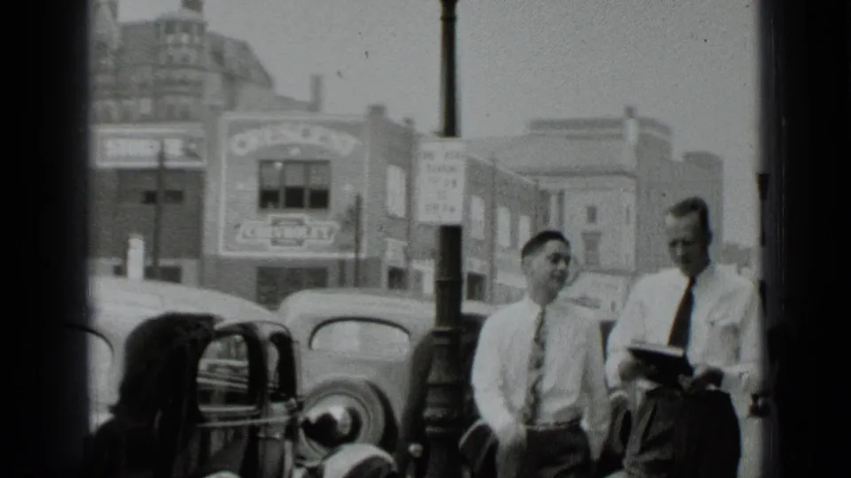 Two men walk past classic cars on a New York city street in 1939 home-movie footage, with commercial signage behind them.