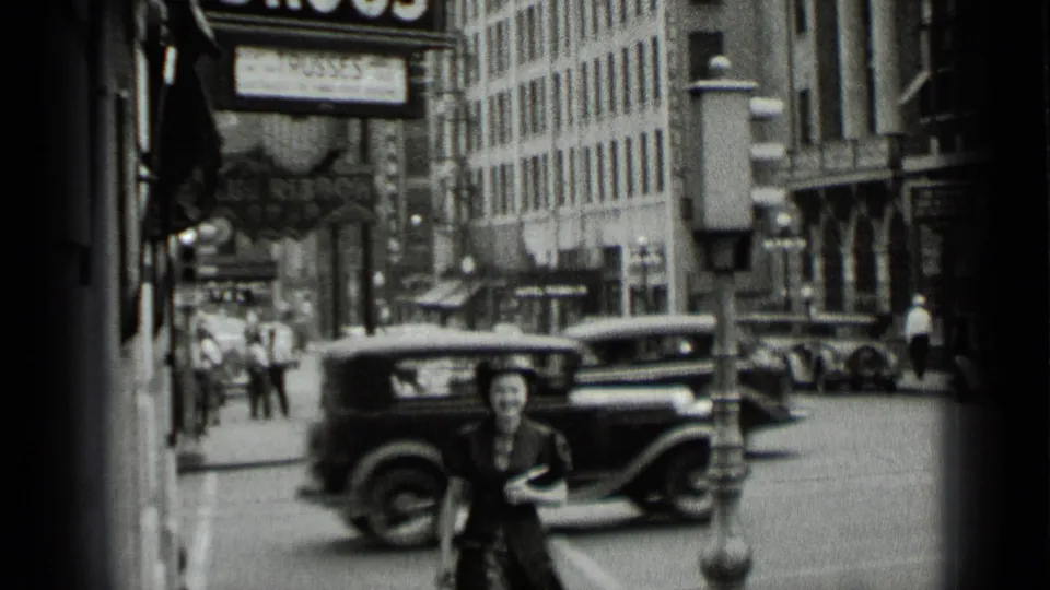 A woman crosses a New York street in 1939 home-movie footage, with moving automobiles and commercial buildings around her.