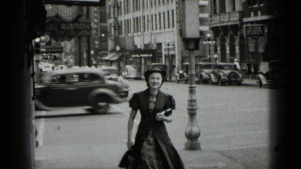 A woman in dark clothing crosses a 1939 New York street while period cars move past commercial buildings in the background.