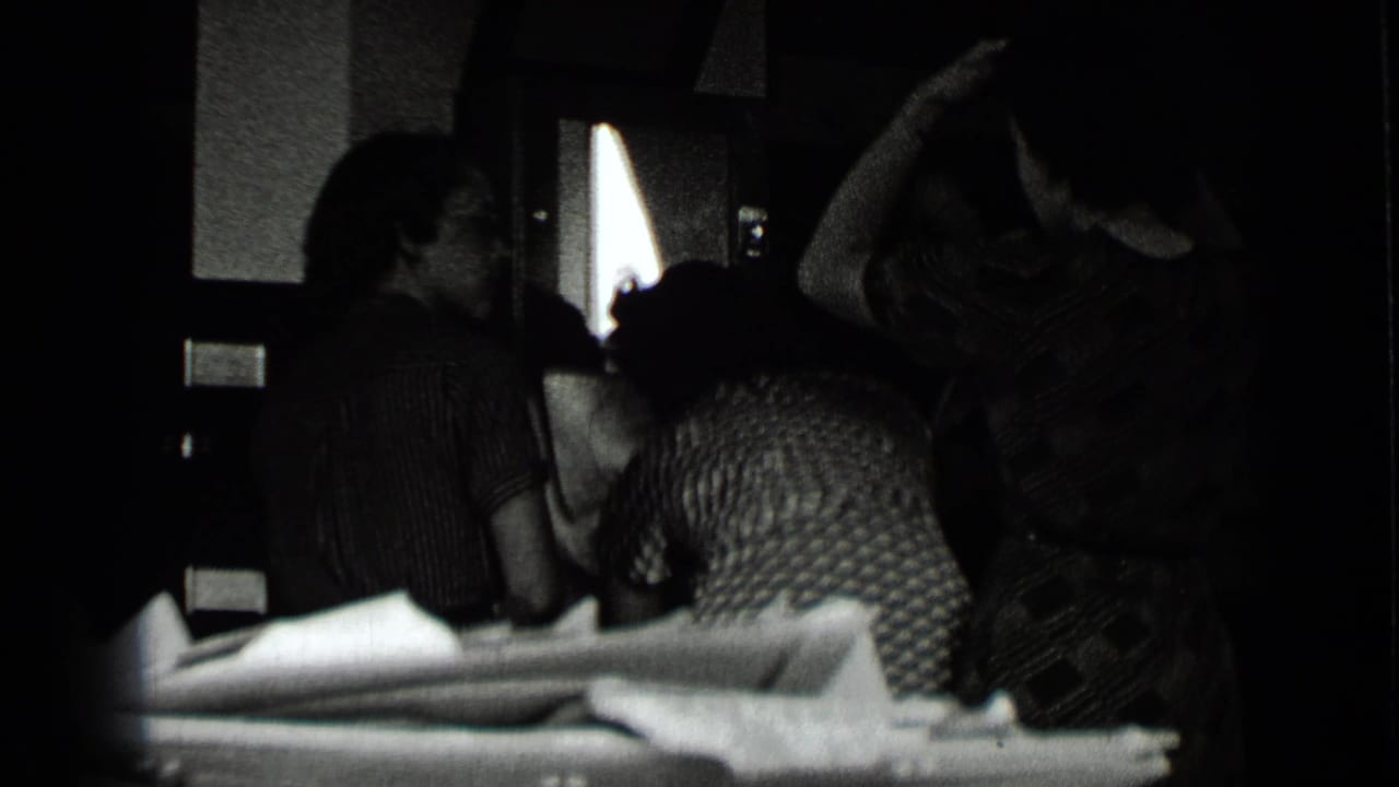 Three women in a dark interior near a bright window, one lifting an arm near her hair.