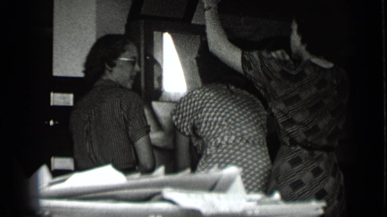 Archival 8mm frame from 1939 New York showing women socializing indoors beside a bright window, with faces partly visible.