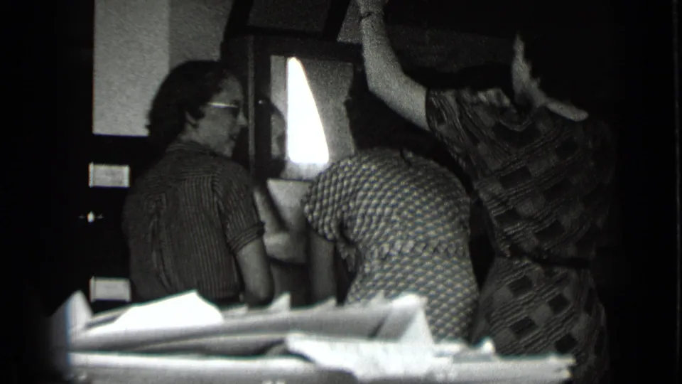 Archival 8mm frame from 1939 New York showing women socializing indoors beside a bright window, with faces partly visible.