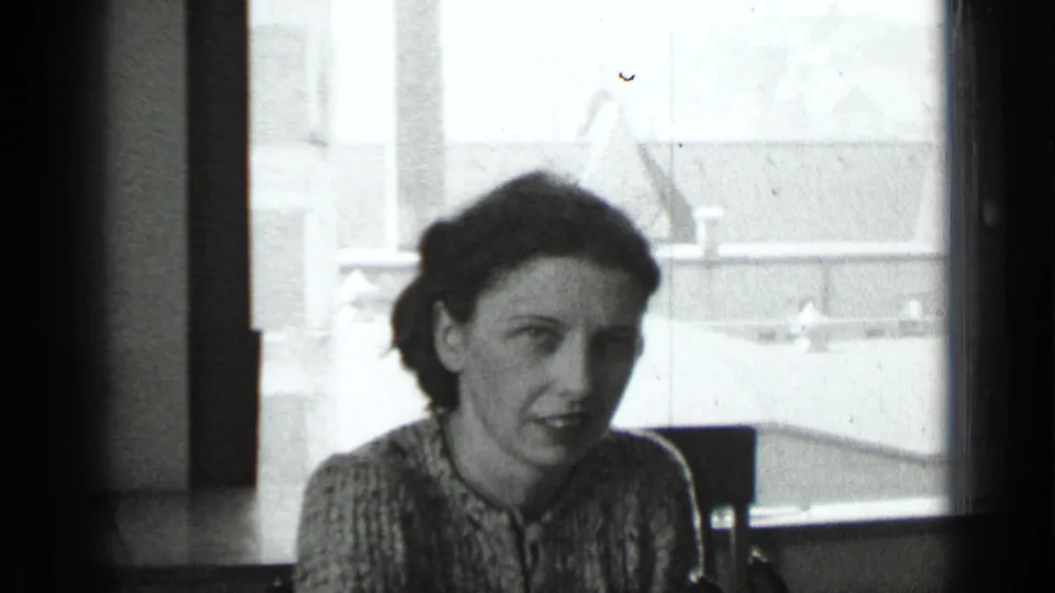 Archival 8mm frame from 1939 New York showing a woman near a bright window, with urban rooftops visible beyond the glass.