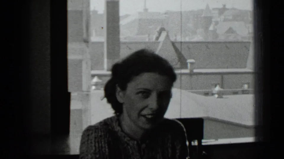Archival 8mm frame from 1939 New York showing a woman smiling or speaking near a bright window with city rooftops behind her.