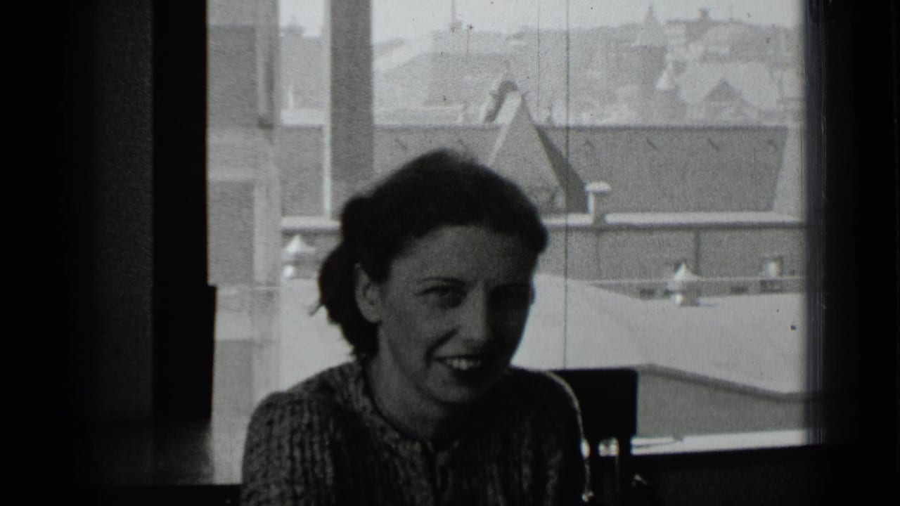 Dark-haired woman smiling near a bright window with city rooftops outside.
