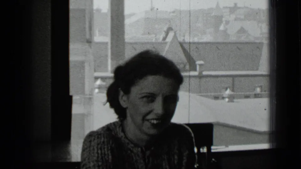 In 1939 New York home-movie footage, a dark-haired woman smiles indoors beside a window overlooking nearby buildings.