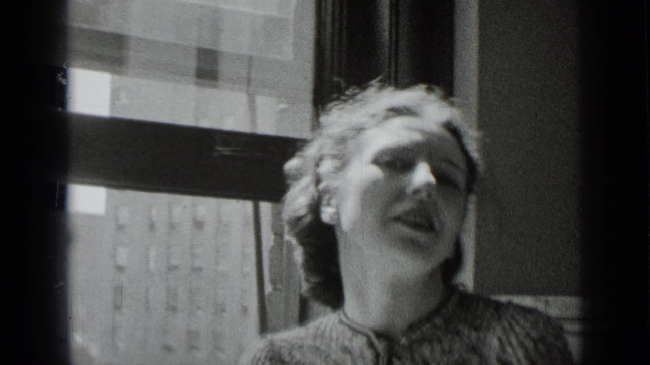 A light-haired woman in 1930s attire appears to speak animatedly near a New York window in black-and-white 8mm footage.
