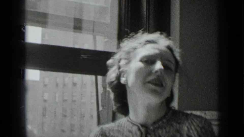 A light-haired woman in 1930s attire appears to speak animatedly near a New York window in black-and-white 8mm footage.