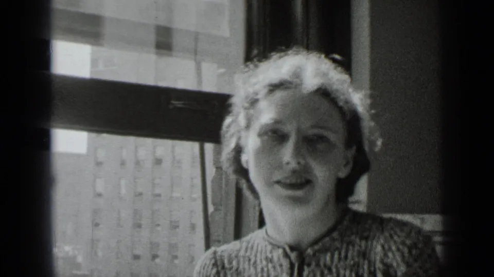 The light-haired woman faces the camera near a sunlit window, with a city building visible through the glass.