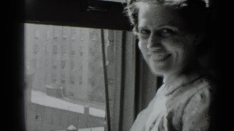 The woman turns slightly and smiles beside the window, framed closely against the urban exterior beyond.