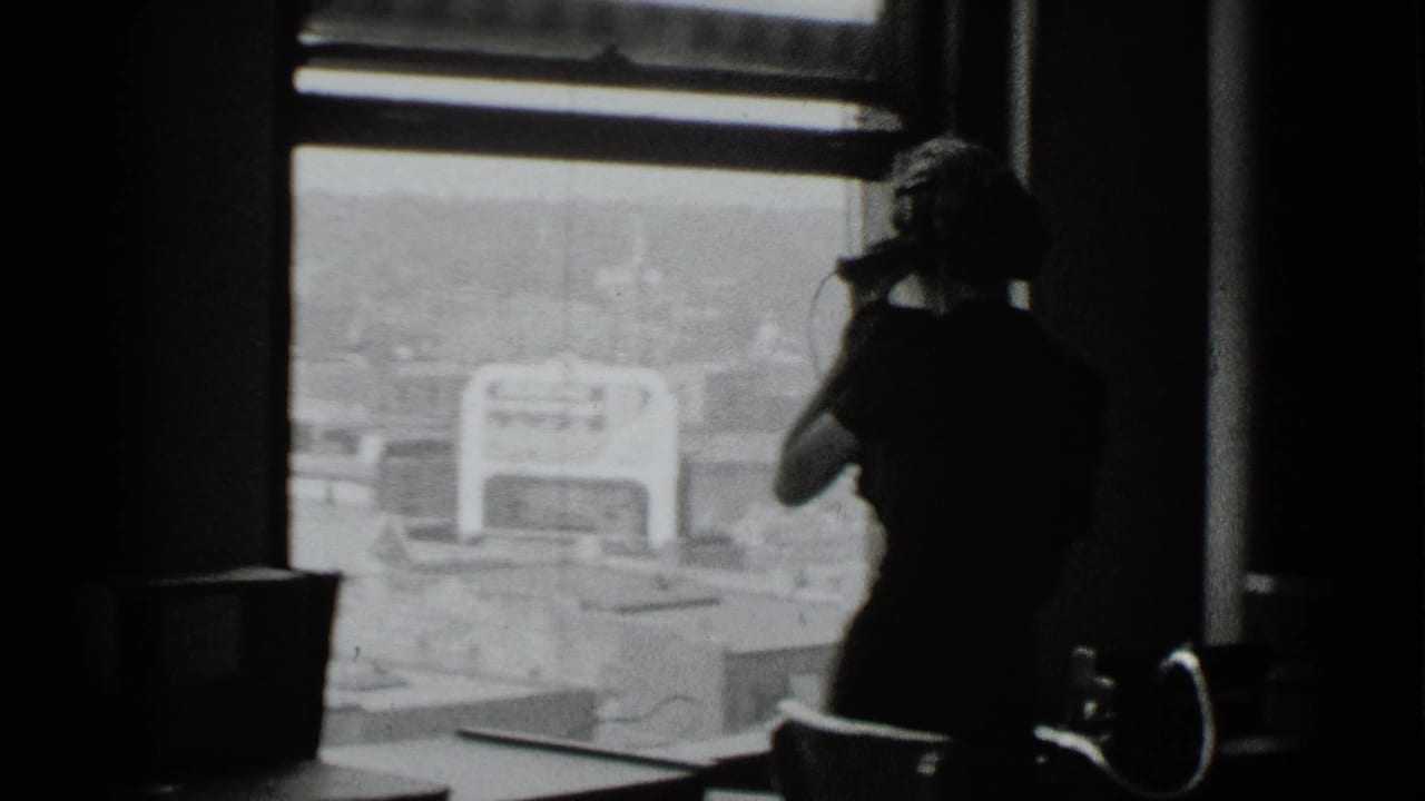 In 1939 New York home-movie footage, a woman stands near a large window with a telephone handset against a bright city view.