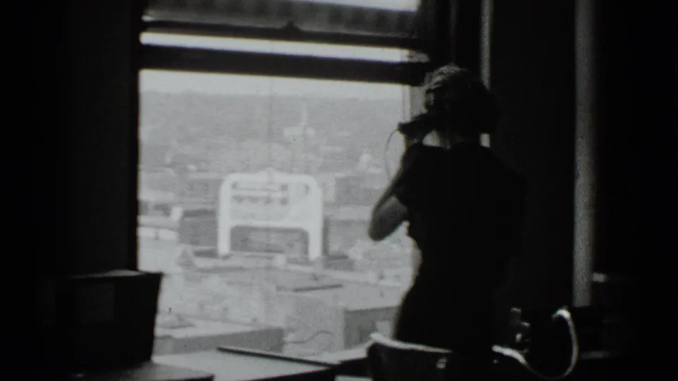 In 1939 New York home-movie footage, a woman stands near a large window with a telephone handset against a bright city view.