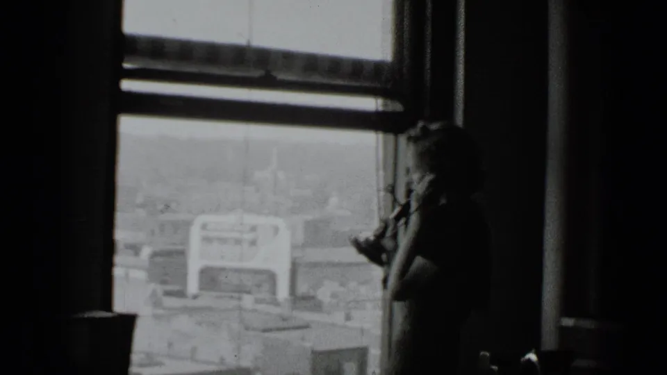 A woman in a dim interior holds a telephone by a tall window, with New York buildings visible outside.