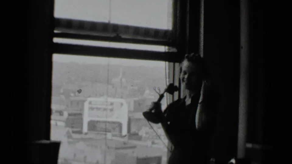 1939 New York home-movie frame showing a dim high-rise interior, large window, city view, and a partly visible woman at left.